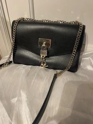 Svart DKNY handväska med guldkedja - Stilren svart handväska från DKNY i äkta läder med grov guldfärgad kedjerem. Väskan har ett metallås framtill och klassisk DKNY-logotyp som hänge. Rektangulär form och elegant design som ger en lyxig känsla. Använd endast en gång sedan inköp. 