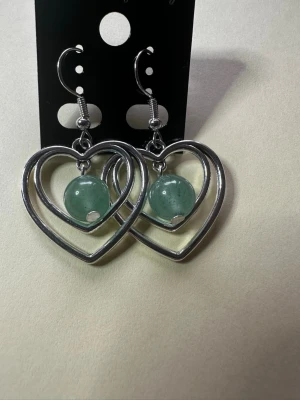 Hjärformade örhängen med grön pärla av aventuriner  - Snygga örhängen i form av silverfärgade hjärtan med en grön pärla i mitten av aventuriner Perfekta för att ge din stil en unik touch. Hängande modell med krokfäste. Passar dig som gillar trendiga accessoarer med lite extra färg.