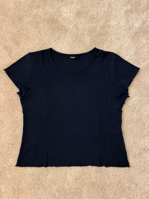 Söt mörkblå topp från ONLY - Söt mörkblå ribbad topp från ONLY med rund halsringning och korta ärmar. Toppen har en croppad passform och är tillverkad i ett mjukt material som sitter bekvämt. Perfekt att matcha med jeans eller kjol. Mindre i storleken.