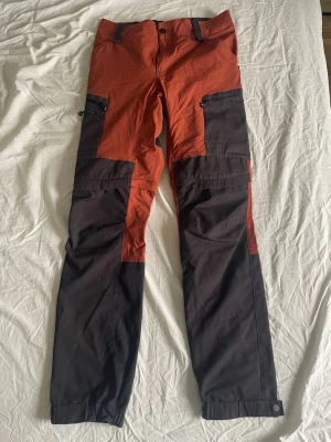 Revolution Race zip off byxor - Funktionella zip-off byxor i röd och mörkgrå färg. Byxorna har dragkedjor vid knäna så de kan göras om till shorts, flera fickor med dragkedja och förstärkta partier. Perfekta för utomhusaktiviteter och äventyr.