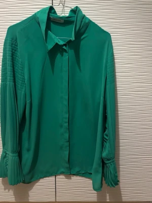 Grön blus med plisserade ärmar Malvin - Stilren grön blus från Malvin med plisserade detaljer på de långa ärmarna och en snygg knytkrage. Blusen har en rak passform och dolda knappar framtill. Perfekt för dig som gillar en trendig och färgstark look.