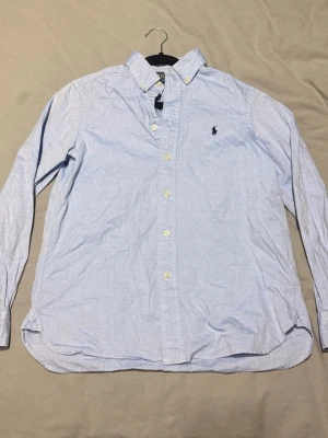 Ljusblå skjorta från Polo Ralph Lauren - Klassisk ljusblå skjorta från Polo Ralph Lauren med broderad logga på bröstet. Skjortan har knappar framtill, button-down krage och långa ärmar. Perfekt för en stilren look.