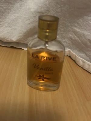 La Rive Vanilla Touch parfym - La Rive Vanilla Touch är en parfym med söt vaniljdoft. Flaskan är genomskinlig med rundade kanter och har ett guldfärgat lock. Texten på flaskan är i guld och brunt, och det finns en liten vaniljblomma på framsidan. Perfekt för dig som gillar varma, mjuka dofter. mer än hälften kvar.