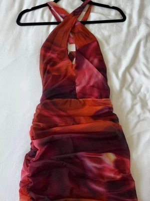 Röd omlottklänning från Oh Polly - Snygg kort klänning från Oh Polly med omlott-design och korsade axelband. Klänningen har ett abstrakt mönster i röd, orange och mörka toner, samt en ringdetalj i ryggen. Materialet är polyester och klänningen är tight med rynkade detaljer.