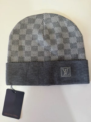 L, V mössa - Snygg grå mössa från L, V med klassiskt rutmönster och uppvikt kant. L, V-logotypen är broderad på framsidan. Mössan är tillverkad i mjukt stickat material och har en modern, stilren look som passar perfekt till streetwear.