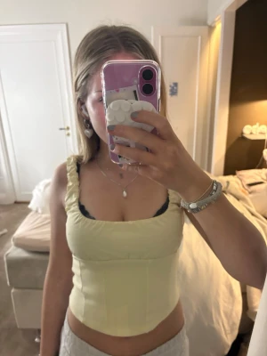 Ljusgul korsett-topp med volang - Säljer en ljusgul korsett-topp med volangdetaljer på axelbanden. Toppen har en tight passform och är ärmlös med breda axelband. Perfekt för dig som vill ha en trendig och feminin look. Materialet känns som bomull.