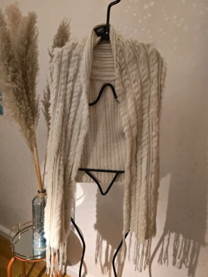 Beige stickad väst med fransar - Mysig beige stickad väst från HOLLIES med långa fransar nertill och ett fluffigt, kabelstickat mönster. Västen är öppen framtill och har en avslappnad passform, perfekt att slänga över en tröja för extra stilpoäng.