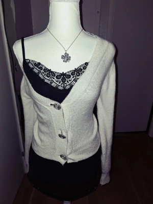 Vit kofta med dekorativa knappar - Mysig vit kofta med långa ärmar och djup V-ringning. Koftan har tre stora dekorativa knappar framtill och är tillverkad i ett mjukt, stickat material. Passar perfekt över ett linne eller topp för en snygg lager-på-lager-look.