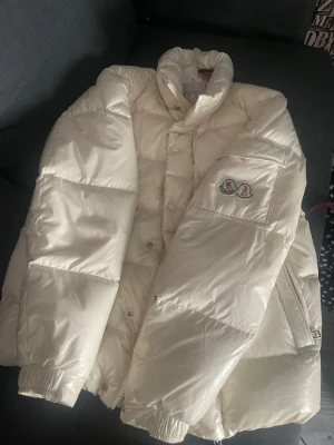 Vit pufferjacka från Moncler - Snygg och fluffig vit pufferjacka från Moncler med hög krage och klassisk logotyp på ärmen. Jackan har breda kanaler, tryckknappar och dragkedja samt elastiska muddar. Perfekt för kalla dagar och riktigt varm. Materialet är glansigt och känns lyxigt.