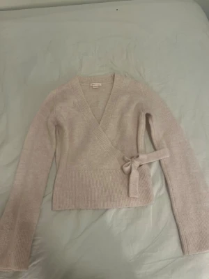 Ljusrosa omlottkofta från Gina Tricot - Supersöt ljusrosa omlottkofta från Gina Tricot med knytband i midjan och ribbade muddar. Koftan har lång ärm och är stickad i ett mjukt material som känns riktigt skönt mot huden. Perfekt för lager-på-lager och en chill look.