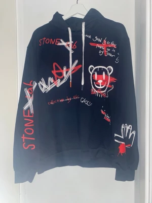 Svart hoodie med grafiska tryck - Svart hoodie med unika grafiska tryck i rött och vitt, inklusive text och olika motiv. Hoodien har huva med vita snören och ribbade muddar. Perfekt för dig som gillar streetwear och vill sticka ut med en kreativ look. 