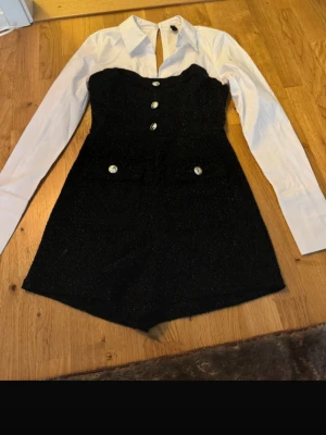 Svart och vit jumpsuit med knappar - Snygg jumpsuit med svart glittrig byxdress och vita långärmade skjortärmar. Framsidan har dekorativa silverfärgade knappar och två fickor. 