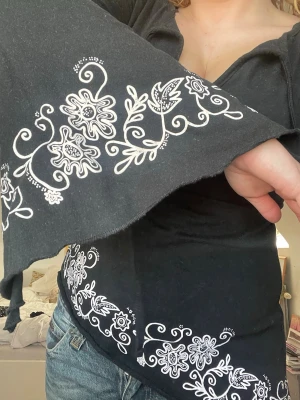 Asymmetrisk topp med vita blommiga detaljer - Svart asymmetrisk långärmad topp med utsvängda ärmar och vita blommiga broderier längs ärmar och nederkant. Toppen har en lätt v-ringad halsringning och sitter snyggt figurnära. Perfekt för dig som gillar boho-stil och unika detaljer.