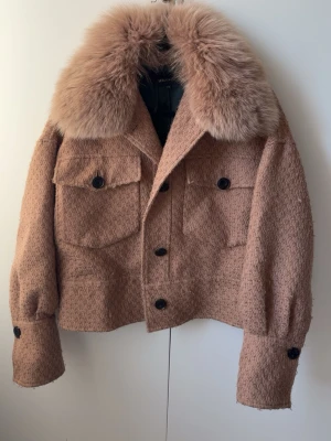 Beige pälsjacka från Meotine - Säljer en beige/brun pälsjacka från Meotine med stor fluffig fuskpälskrage och grovstickad struktur. Jackan har två stora bröstfickor med lock och svarta knappar framtill samt vid ärmsluten. Perfekt för lite kallare vårdagar 