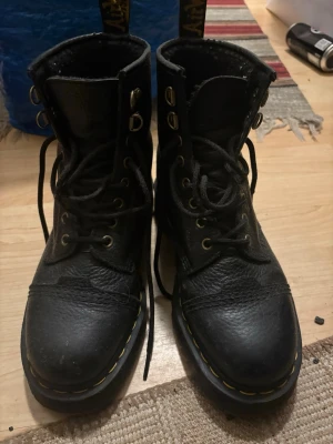 Svarta Dr. Martens kängor i läder - Säljer ett par klassiska svarta Dr. Martens kängor i grovt läder med gul söm runt sulan. Högt skaft, snörning med metallhål och krokar. Perfekta för dig som gillar en edgy och tidlös stil.