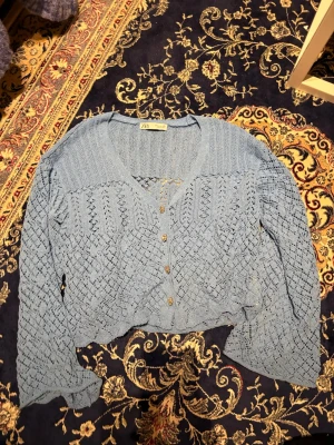 Blå stickad kofta från Zara - Säljer en ljusblå stickad kofta från Zara med v-ringning och dekorativa guldknappar framtill. Koftan har ett genombrutet mönster och långa ärmar. Perfekt att slänga över en topp för en trendig lager-på-lager-look.