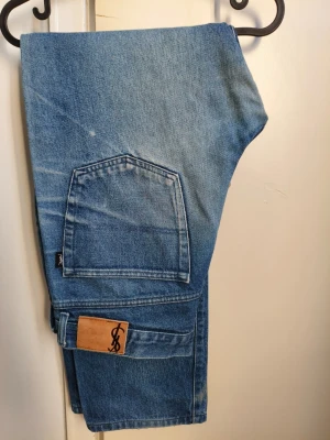 Blå vintagejeans från Yves Saint Laurent - Säljer ett par klassiska blå jeans från Yves Saint Laurent med raka ben. Jeansen har stora bakfickor, YSL-läderpatch bak i midjan och är gjorda i slitstark denim. Perfekta för dig som gillar retrostil och tidlös design.