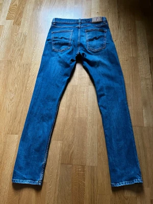 Nudie Jeans Lean Dean - Klassiska blå jeans med rak passform och fem fickor. Jeansen har normalhög midja och subtila slitningar framtill. Perfekta för en avslappnad och stilren look. Materialet är denim i bomull med lätt stretch för extra komfort.