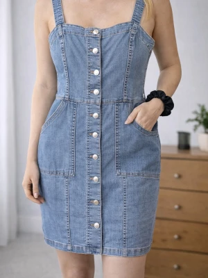 Jeansklänning med knappar framtill - Snygg jeansklänning i blå denim med breda axelband och knappar längs hela framsidan. Klänningen har två stora fickor framtill och markerad midja för en smickrande passform. Perfekt för dig som gillar en avslappnad men trendig stil.
