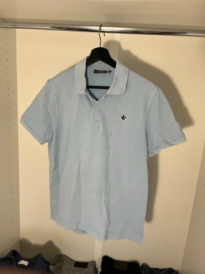 Ljusblå pikéskjorta från Morris - Ljusblå pikétröja från Morris med klassisk krage, två knappar och broderad logga på bröstet. Tillverkad i mjuk bomull och har korta ärmar. Hör av dig vid frågor!