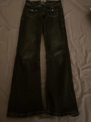 Svarta bootcut jeans med fickdetaljer - Säljer ett par svarta bootcut jeans med snygga fickor framtill och kontrastsömmar. Jeansen har knappar på ficklocken och är ifrån Gina Young alltså Gina tricot barn. Perfekta för dig som gillar en avslappnad men ändå trendig stil.