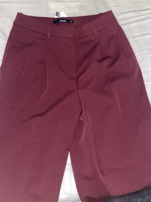 Vinröda kostymbyxor från Bik Bok - Snygga vinröda kostymbyxor från Bik Bok med raka ben och pressveck. Byxorna har hällor för skärp, två sidofickor och stängs med dragkedja och hake. Perfekta för en stilren och trendig look.