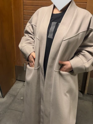 Beige lång kappa med fickor - Stilren beige kappa i lång modell med bred krage och två stora fickor framtill. Kappan har en avslappnad passform och är tillverkad i ett mjukt, tjockt material som ger en lyxig känsla. Perfekt för dig som gillar minimalistisk och modern stil.