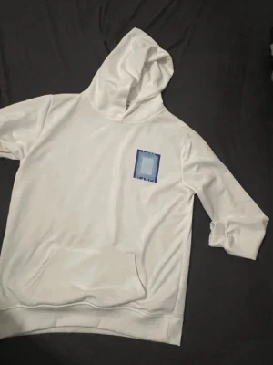 Vit hoodie från Casablanca med blått tryck - Snygg vit hoodie från Casablanca med blått grafiskt tryck på bröstet och ryggen där det står 'Casa Blanca'. Hoodien har huva, magficka och ribbade muddar. Perfekt för en chill och stilren look.