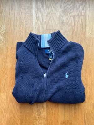 Mörkblå stickad ziptröja Polo Ralph Lauren - Mörkblå stickad ziptröja från Polo Ralph Lauren med ljusblå broderad logga på bröstet. Hög ribbad krage med ljusblå och vita ränder på insidan. Perfekt för lager-på-lager och snygg till jeans och linnebyxor. Storleken är 14-16.
