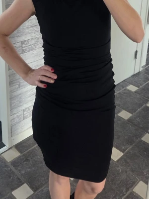 Svart ärmlös kortklänning - En stilren svart klänning med tight passform och ärmlös design. Klänningen har rynkade detaljer runt midjan som ger en snygg siluett. Perfekt för dig som gillar enkel och klassisk stil. Materialet känns mjukt och stretchigt, vilket gör den bekväm att bära. Bestäm längden själv. 