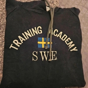 Hoodie Training Academy SWE M - Svart hoodie Training Academy SWE  Svart hoodie från Interco Classic Wear i storlek M med broderad text 'Training Academy SWE' och svensk flagga på bröstet. Tröjan har huva med snörning, känguruficka och ribbade muddar. Insidan av huvan är ljus och materialet är mjuk bomull.  Kan även hämtas I Partille  E1
