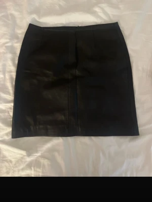 Svart kort skinnkjol WERA - Snygg svart kort skinnkjol med stilren design och raka linjer. Kjolen har en klassisk passform och dragkedja bak. Kjolens sömm har gått upp där bak (se bild).