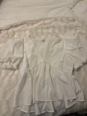 Vit puffärmad omlottblus - Snygg vit omlottblus med puffiga korta ärmar och markerad midja. Blusen har ett flowy fall och är tillverkad i ett tunt, lätt tyg som ger en luftig känsla. Perfekt att styla med jeans eller kjol för en trendig look. Den har en liten foundation fläck uppe på axeln .
