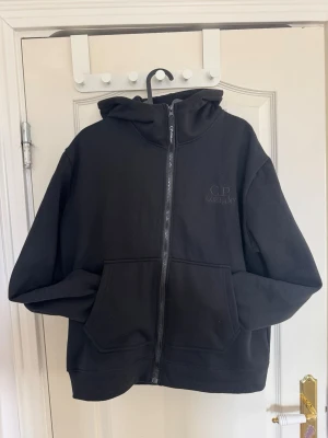 Svart hoodie med glasögon från C.P. Company - Svart hoodie med dragkedja från C.P. Company. Klassisk modell med två stora fickor fram och huva med inbyggda goggles. Broderad logga på bröstet och mjukt bomullsmaterial. Perfekt för en streetwear-look. Ej blivit använd pga att den är för liten för mig. Säljer derför den vidare nu. Hör av er vid minsta fråga :) 