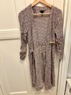 Blommig omlottklänning med knytband 🌸 - Jättefin omlottklänning med blommigt mönster i rosa, vit och svart i 70-talsstil. Klänningen har knytband i midjan och en avslappnad passform. Perfekt för dig som gillar en feminin och bekväm stil. Bra skick förutom att en knapp i midjan har gått av vilket åtgärdats med säkerhetsnål. 🌸