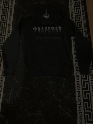 Svart hoodie från Trapstar - Snygg svart hoodie från Trapstar med stor logga och text på bröstet. Klassisk huva och magficka framtill. Perfekt för en streetwear-look och passar till det mesta i garderoben.