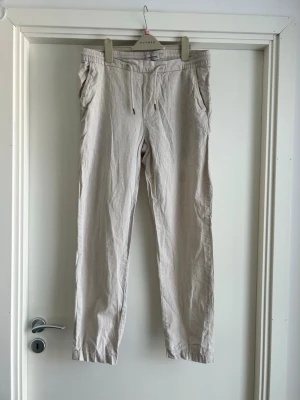 Beige linnebyxor Dressmann - Snygga beige linnebyxor från Dressmann med avslappnad passform. Byxorna har sidfickor fram och knappar bak. Perfekta för en chill och stilren look under varma dagar. Tillverkade i ett lätt och luftigt linnematerial.