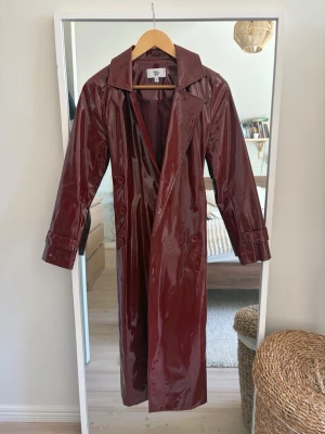 Vinröd lack trenchcoat från Rebecca Stella - Säljer en vinröd trenchcoat i glansigt lackmaterial från Rebecca Stella. Jackan har dubbelknäppning, bred krage och långa ärmar. 