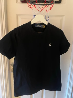 Svart t-shirt från Polo Ralph Lauren - Klassisk svart t-shirt från Polo Ralph Lauren med den ikoniska vita logon broderad på bröstet. T-shirten har rund hals och korta ärmar, tillverkad i mjuk bomull för en skön känsla. Perfekt basplagg för alla tillfällen.