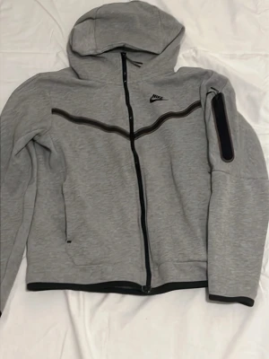 Grå hoodie från Nike med dragkedja - Snygg grå hoodie från Nike med hel dragkedja och svart detalj längs bröstet. Huva och praktisk ficka på ärmen. Perfekt för en sportig och avslappnad stil.