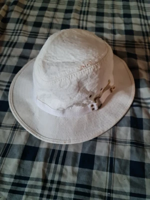 Vit bucket hat i bomull - Snygg vit bucket hat i bomull med bred brätte och justerbart band på sidan. Perfekt för soliga dagar och ger en avslappnad streetstyle-look. Hatten har metallspännen och klassisk form som passar till många outfits.