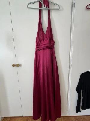 Vinröd/rosa balklänning i satin - Elegant vinröd/rosa långklänning i glansigt satinmaterial med halterneck och djup v-ringning. Klänningen har draperad midja. Storlek 34 och nypris 1200kr. Lite småskavanker längst ner men inget man lägger märke till. Otroligt fin banklänning 🫶