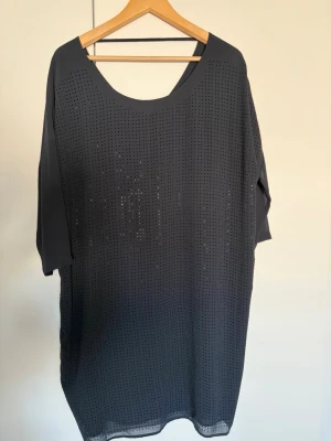 Svart loose klänning med hålmönster - Mörkblå klänning med loose passform och trekvartsärm. Framsidan har skimrande stenar som ger en cool och modern look. Klänningen har rund halsringning och är tillverkad i ett lätt syntetmaterial. Perfekt för dig som gillar enkel och stilren design. Nypris 699kr