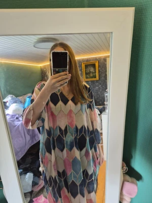 Mönstrad kortklänning med v-ringning - Säljer en färgglad kortklänning med v-ringning och trekvartsärm. Klänningen har ett geometriskt mönster i blått, rosa, beige och vitt. Materialet känns mjukt och luftigt, perfekt för varmare dagar. Passformen är loose och klänningen har en avslappnad stil.