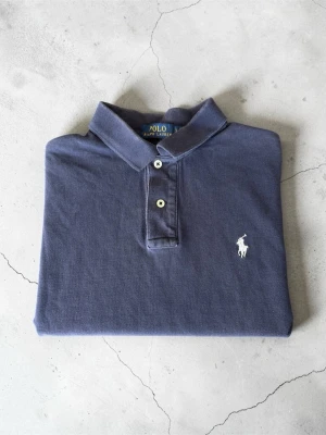 Ralph Lauren Piké – Mörkblå | Klassisk & Tidlös - Klassisk marinblå pikétröja från Polo Ralph Lauren med vit broderad logga på bröstet. Tröjan har korta ärmar, krage och två knappar framtill. Tillverkad i mjuk bomull som känns skön mot huden. Perfekt för en clean och stilren look.