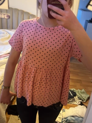 Rosa prickig tröja med volang - Supersöt rosa blus med svarta prickar och volang nedtill. Blusen har korta ärmar och en lös passform som ger en avslappnad vibe. Perfekt att styla med jeans eller kjol för en trendig look. Tröjan är lite noppig men inget som förstör.