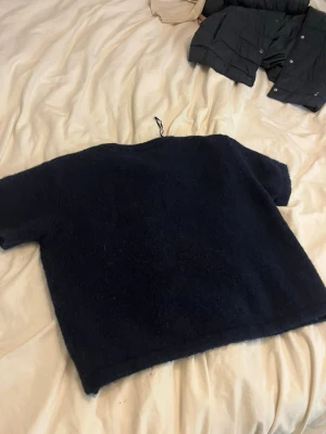 Mörkblå stickad tröja Gina Tricot - Mysig mörkblå stickad tröja från Gina Tricot med korta ärmar och rund halsringning. Tröjan har en fluffig struktur och ribbstickade detaljer vid halsen. Perfekt för lager-på-lager och enkel att matcha med jeans eller kjol.