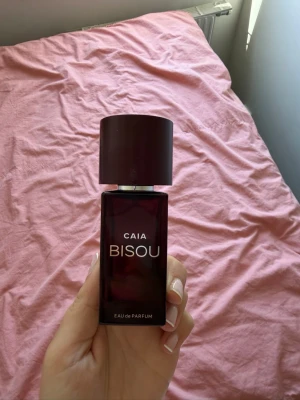 CAIA Bisou Eau de Parfum - Säljer en CAIA Bisou Eau de Parfum i en snygg mörkröd, fyrkantig glasflaska med matchande lock. Flaskan har en modern och stilren design som känns lyxig och trendig. Perfekt för dig som vill ha en unik och elegant parfym i samlingen. Lite mer är halva kvar 