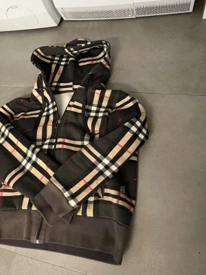 Rutig hoodie med dragkedja från burberry - Snygg hoodie med klassiskt rutigt mönster i beige, svart, vitt och rött. Dragkedja framtill, huva och muddar vid ärmslut och nederkant. Insidan är mjuk och fodrad för extra komfort. Perfekt för en trendig och avslappnad stil. Väldigt grich 