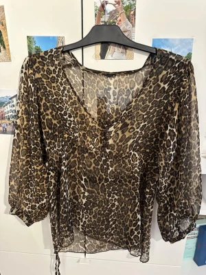 Leopardmönstrad transparent blus - Snygg blus i transparent material med leopardmönster. Blusen har v-ringning, trekvartsärmar med lätt puff och knytband i midjan. Perfekt för dig som vill sticka ut med ett djurmönstrat plagg. Materialet är tunt och luftigt.